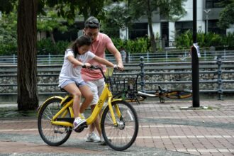 Roles Of The Father In The Family: Father teaches daughter to ride a yellow bicycle in a Singapore park. Bonding moment.