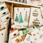 Introduction to Pattern and Card Making: Artistic holiday card designs with watercolor paints and brushes on a wooden table.