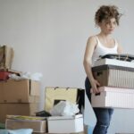 Uses Of Personal And Family Resources: A young woman carrying boxes during a move into a new apartment, surrounded by packed belongings.