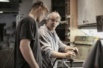 Apprenticeship: Side view of senior foreman in eyeglasses showing to trainee how handling detail in workshop