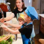 Roles Of The Children In The Family: Mother and children wearing chef hats washing hands while preparing food together in the kitchen.