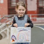 introduction to drawing - A cheerful child proudly shows her rainbow drawing while standing in a playground.