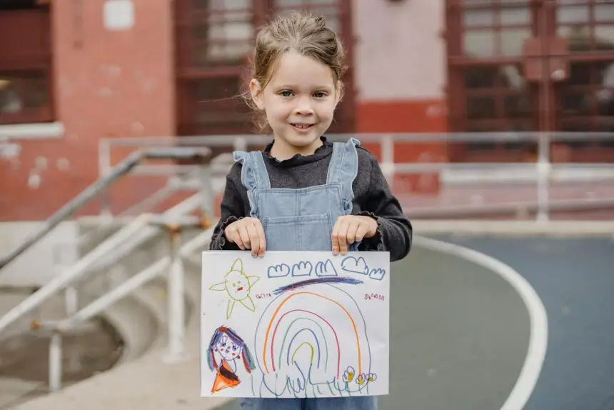 introduction to drawing - A cheerful child proudly shows her rainbow drawing while standing in a playground.